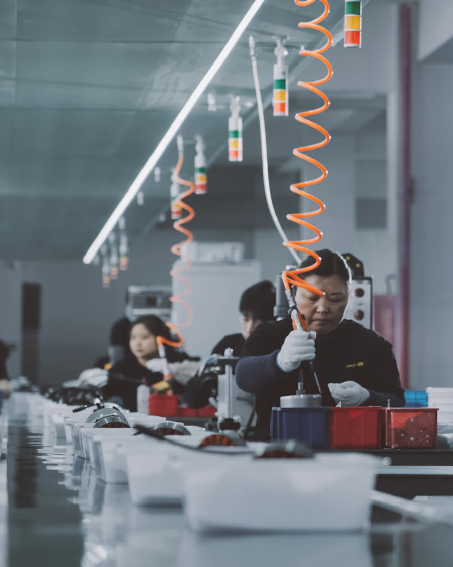 Workers at a bench assembly line beneath coiled air hoses.